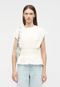 Mujer con blusa blanca sin mangas con cintura fruncida y jeans azul claro, llevando un bolso de hombro plateado con detalles de flecos.