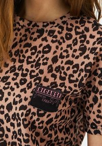 Tričko se vzorem leopardí kůže v světle růžové barvě s černými skvrnami. Má černou náprsní kapsu s bílým nápisem „PROTOŽE VÍŠ, ŽE JSEM.“ Klasický kulatý výstřih.