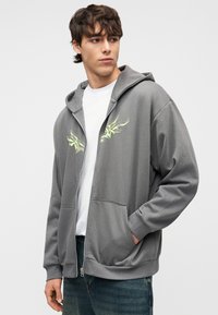 Νέος άνδρας που φοράει γκρι φούτερ με φερμουάρ και κίτρινες αφηρημένες σχεδίασεις, λευκό T-shirt από μέσα και μπλε τζιν, ποζάροντας μπροστά σε ένα απλό φόντο.