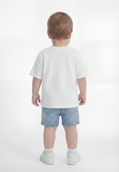 Παιδί με ανοιχτά καστανά μαλλιά, ντυμένο με λευκό μπλουζάκι, μπλε τζιν σορτς, λευκές κάλτσες και λευκά αθλητικά παπούτσια, στέκεται γυρισμένο με την πλάτη σε λευκό φόντο.