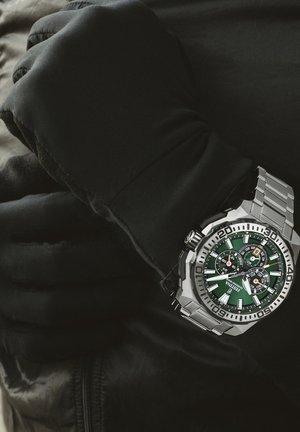 Orologio in acciaio inox argentato con quadrante verde, tre sottomisure e una lunetta unidirezionale. Il polso è coperto da un guanto nero.