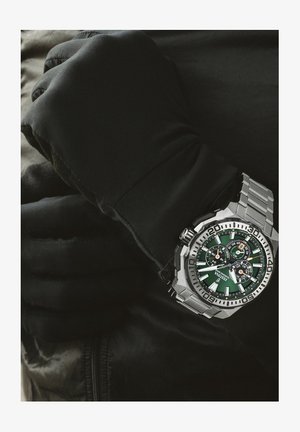 Orologio in acciaio inox argentato con quadrante verde, tre sottomisure e una lunetta unidirezionale. Il polso è coperto da un guanto nero.