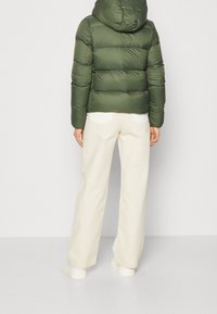 Zielona pikowana kurtka z kapturem i poziomym przeszyciem, w połączeniu z kremowymi szerokimi spodniami i białymi trampkami, widziana od tyłu.