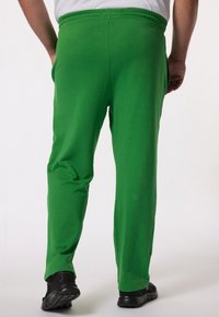 Groene sportbroek van zacht materiaal, met een elastische tailleband, zijzakken en een losse pasvorm die aan de enkels taps toeloopt. Zwarte schoenen zichtbaar.