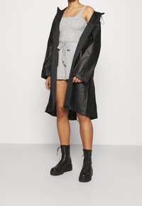 Zwarte waterdichte lange jas over een geruite romper met taillestrik, gecombineerd met zwarte veterlaarsjes. Gladde en textuurrijke materialen.