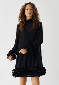 Črna obleka z dolgimi rokavi, ki ima nabrane dodatke na manšetah in robu. Narejena iz lahkega materiala z prozornim prekrivanjem.