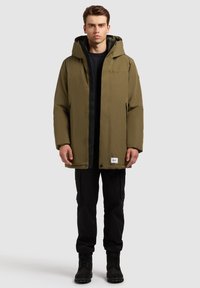 Olivengrøn, vandtæt parka med hætte, frontlukning med trykknapper og sidelommer, båret over en sort sweatshirt og cargobukser. Sorte støvler.