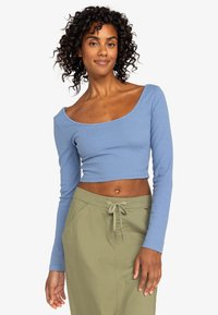 Blå langærmet ribbet crop top med vid scoop neckline, parret med olivengrønne bukser med snøre og sidelommer.