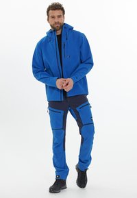 Sininen pehmeäkuorinen takki ja housut, joissa on vetoketjuja, taskuja ja huppu, valmistettu kevyestä, vettä hylkivästä materiaalista; kapea istuvuus.