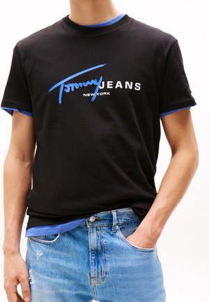 Homme portant un t-shirt noir Tommy Jeans New York par-dessus une chemise bleue, avec un jean bleu clair déchiré et une main dans la poche.
