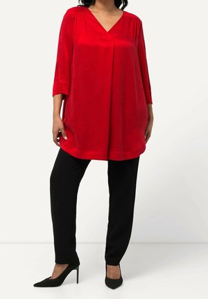 Femme portant une tunique rouge ample avec des manches trois-quarts, un pantalon slim noir et des talons hauts pointus noirs sur fond blanc.