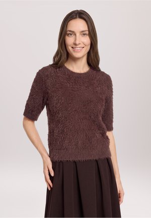 Kvinde smilende, iført en blød brun kortærmet sweater og en mørkebrun plisseret nederdel mod en lys grå baggrund.