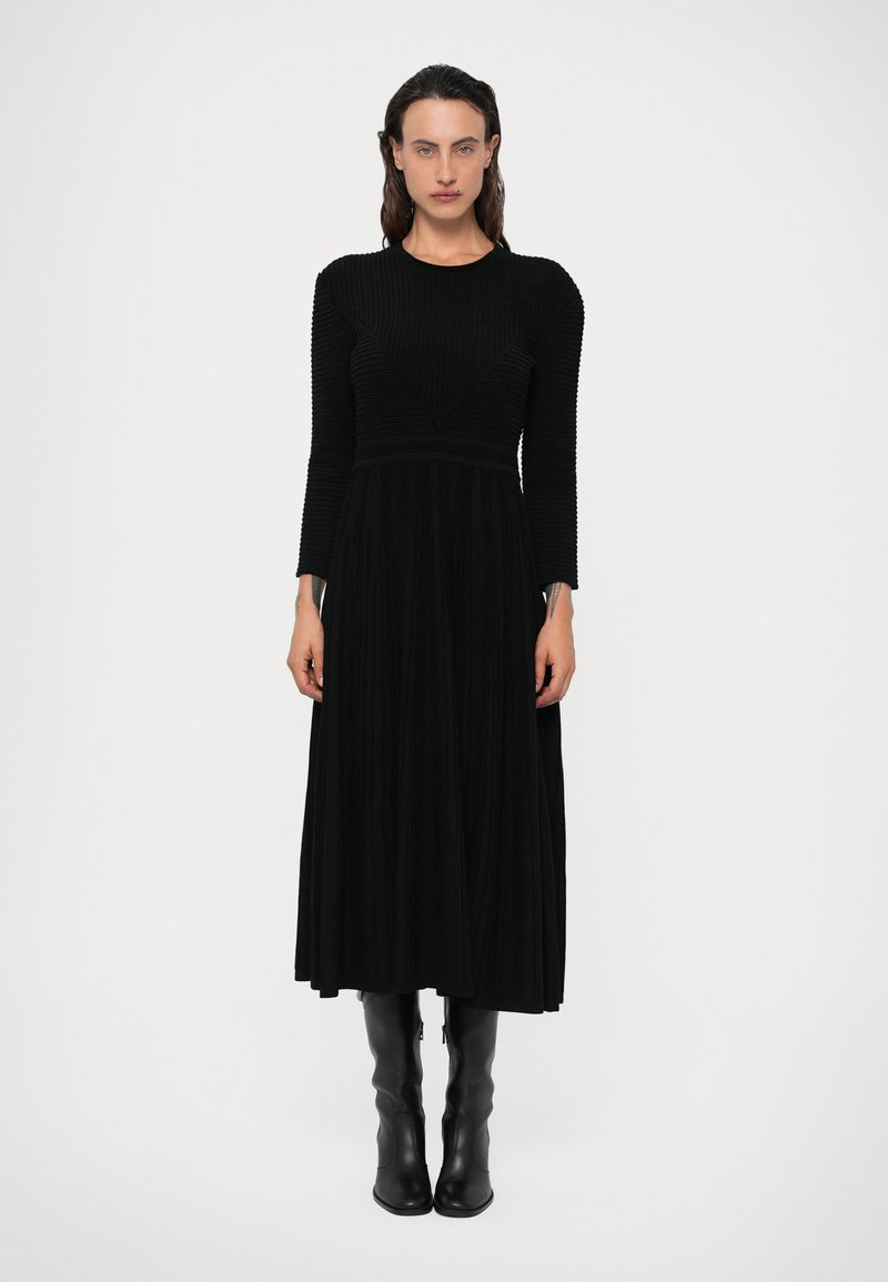 Vestido preto de mangas longas com corpete ajustado, textura canelada e saia fluida e plissada. Combinado com botas pretas até ao joelho.