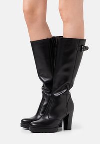 Black knee-high boots made of glossy leather, featuring a side zipper, a buckle accent, and a chunky high heel with textured sole.