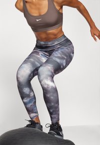 Woman wearing patterned leggings and brown sports bra balancing on a black balance trainer while exercising.