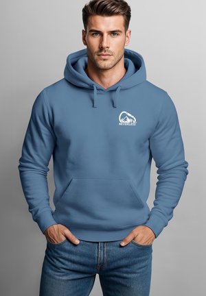 Mann trägt blaue Kapuzenjacke mit Vordertasche und "Neverless" Logo mit Berg und Karabiner, steht vor einem schlichten grauen Hintergrund.