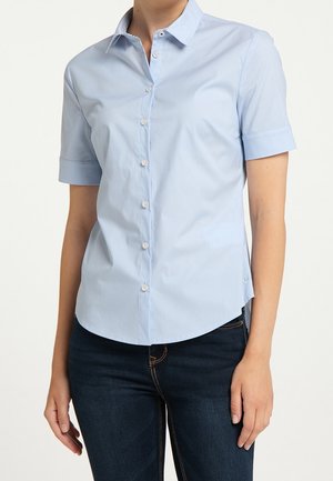 Kobieta ubrana w jasnoniebieską koszulę na krótki rękaw na guziki, zestawioną z ciemnoniebieskimi dżinsami na tle jednolitego tła.