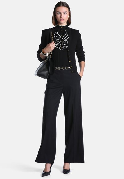 Femme debout portant un cardigan noir, un top noir avec un motif blanc, un pantalon large noir, des talons noirs, des accessoires dorés et portant un sac à main noir.