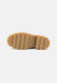Semelle d'une chaussure marron clair avec de profondes rainures horizontales larges pour l'adhérence, présentée sur un fond clair uni.