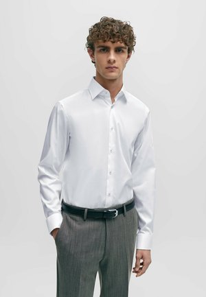 Chemise blanche à manches longues avec un col formel et un devant boutonné, accompagnée d'un pantalon gris à rayures et d'une ceinture noire. Texture lisse.