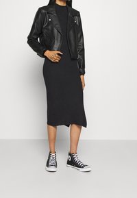 Personne portant une veste en cuir noire, une robe midi côtelée noire et des baskets montantes noires, debout devant un fond clair uni.