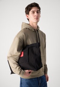 Saco preto de lona com um detalhe vermelho, formato retangular, textura durável; o modelo veste um hoodie bege e jeans azuis, segurando o saco.