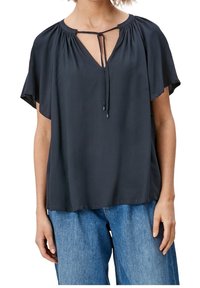 Haut noir à manches courtes avec une finition texturée, présentant un décolleté en V et un détail à nouer au niveau de la poitrine. Associé à un jean en denim bleu clair.