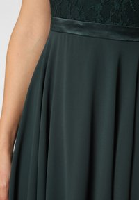 Dark green dress featuring a lace bodice, satin waistband, and flowing fabric with soft pleats. Smooth texture with a classic silhouette.