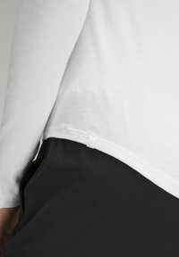 Maglietta bianca a maniche lunghe con orlo arrotondato, abbinata a pantaloni neri in tessuto. Presenta un piccolo etichetta bianca sul lato.