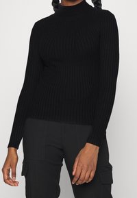 Svart ribbad polotröja med långa ärmar; har en figurnära design och texturerad tyg, ihop med svarta cargobyxor.