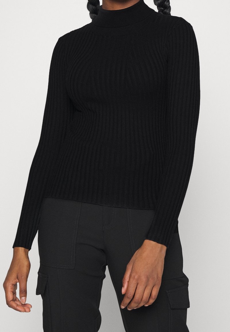 Svart ribbad polotröja med långa ärmar; har en figurnära design och texturerad tyg, ihop med svarta cargobyxor.