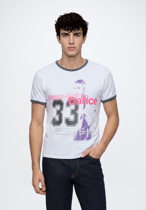 Jeune homme aux cheveux bouclés portant un t-shirt blanc avec bord gris et texte graphique "33 Dance", posant devant un fond uni.