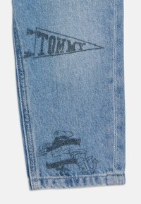 Jeans in denim blu chiaro con grafica nera stampata: un logo triangolare con la scritta "TOMMY" e un design stilizzato vicino all'orlo.