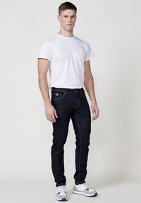 Dunkelblaue Jeans aus Denim mit heller Naht, schmal geschnittenes Design. Kombiniert mit einem schlichten weißen T-Shirt und grauen Sportschuhen. Einfacher, funktionaler Stil.