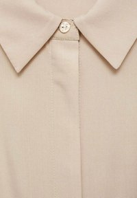 Camicia beige con colletto, caratterizzata da una chiusura con un unico bottone e un tessuto liscio e texture, con cuciture ordinate lungo la parte anteriore.