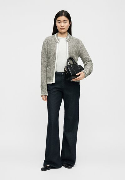 Γυναίκα που στέκεται φορώντας γκρι υφαντό σακάκι, λευκό τοπ, μαύρο παντελόνι φαρδύ, μαύρα παπούτσια, κρατώντας μικρό μαύρο τσαντάκι σε λευκό φόντο.