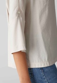 Helle beige Bluse mit lockerer Passform, 3/4-langen Ärmeln, Knopfdetail am Ärmelbündchen, aus einem weichen, strukturierten Stoff gefertigt.