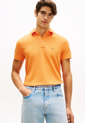 Joven con cabello corto y castaño, vistiendo una camiseta polo naranja y jeans azul claro, de pie con una mano en el bolsillo.