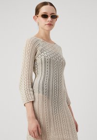Beige, kötött ruha csipkeszerű mintával. Háromnegyedes ujja van, kerek nyakkivágása és olyan fazonja, amely kiemeli az alakot.