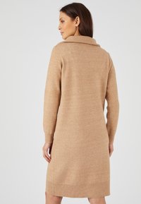 Abito in maglia di colore beige morbido con un ampio colletto a risvolto, maniche lunghe e polsini e bordo a coste. Texture liscia e vestibilità morbida.