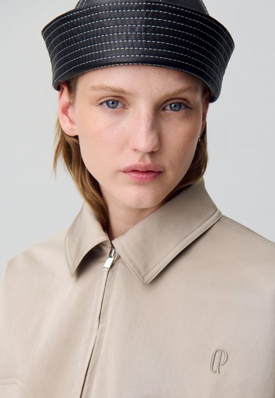 Giacca in pelle beige con colletto e zip argento. Cappello nero testurizzato con cuciture bianche. Dettaglio logo sottile sulla giacca.