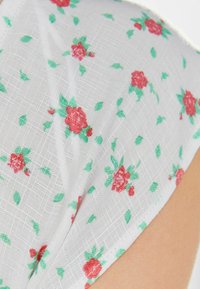 Valkoinen kangas, jossa on teksturoitu kuvio pienistä punaisista ruusuista ja vihreistä lehdistä, antaen pehmeän ja kevyen ilmeen.