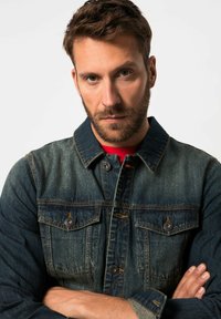 Chaqueta de mezclilla azul con dos bolsillos en el pecho, cierres de botones y costuras marrones. La textura parece resistente con un corte clásico y cuello.