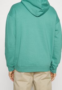 Un hoodie verde con una texture morbida e un grande cappuccio; presenta una vestibilità ampia e polsini e fondo a costine. Indossato sopra pantaloni beige.