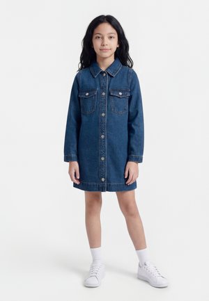 Fată tânără în picioare, purtând o rochie din denim albastru închis cu nasturi, șosete albe și adidași albi, pe un fundal alb simplu.