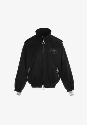 Joven con chaqueta y pantalones negros de gran tamaño con rayas reflectantes plateadas y zapatos negros gruesos, de pie frente a un fondo blanco liso.
