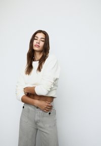 Bela skrajšana trenirka z rebrastimi manšetami, v kombinaciji z svetlo sivo širokimi hlačami z velikimi žepi in kontrastnimi črnimi gumbi.