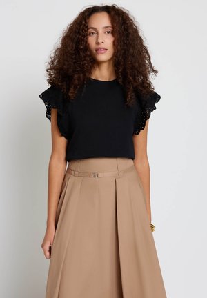 Donna con capelli ricci che indossa una maglia nera con maniche arricciate e una gonna beige a vita alta con cintura, in piedi contro uno sfondo bianco.