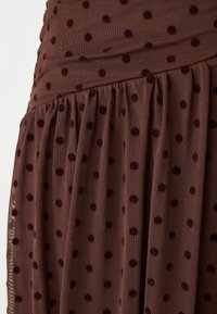 Bruine rok met stippen in een zachte stof en pluche, donkere kastanjebruine stippen. De taille heeft een geraffineerde plooidetail, wat de vloeiende silhoutte benadrukt.