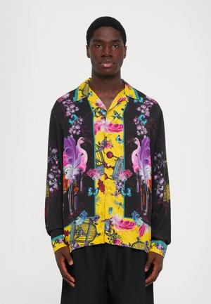 Man wearing a vibrant long-sleeve shirt with yellow, black, pink flamingos, flowers, butterflies, and birdcage patterns, standing against white background.